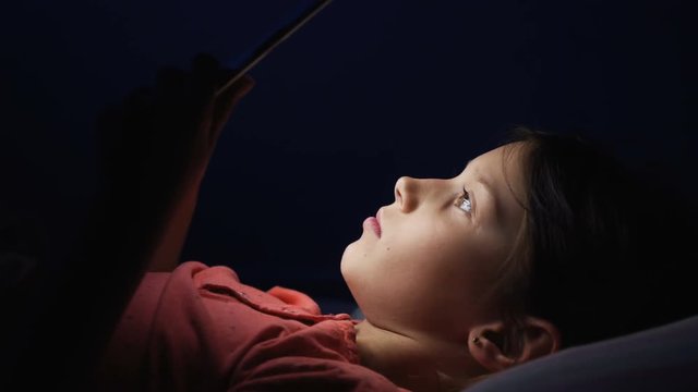 Dolly shot caucasian girl lying in bed playing tablet in social internet in dark blue light under blacket. Childhood, communication and technology concept