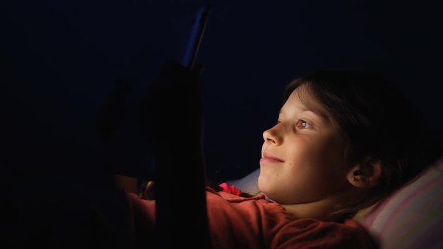 Dolly shot caucasian girl lying in bed playing tablet in social internet in dark blue light under blacket. Childhood, communication and technology concept