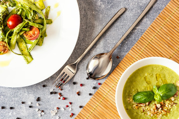 salad with asparagus and cherry tomatoes, green cream soup, silver spoon and fork, grey background