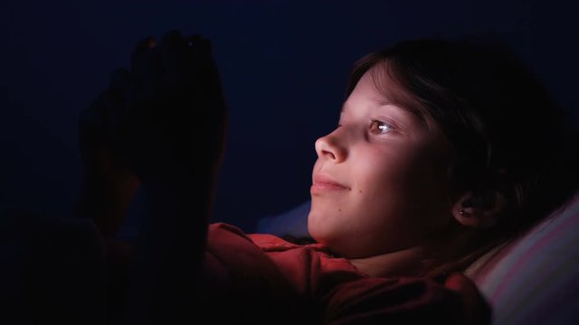 Dolly shot caucasian girl lying in bed playing tablet in social internet in dark blue light under blacket. Childhood, communication and technology concept