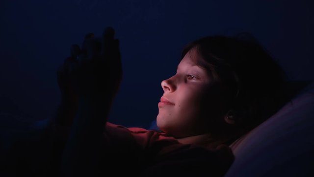 Dolly shot caucasian girl lying in bed playing tablet in social internet in dark blue light under blacket. Childhood, communication and technology concept