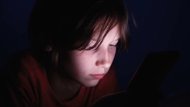 Dolly shot caucasian girl lying in bed playing tablet in social internet in dark blue light under blacket. Childhood, communication and technology concept