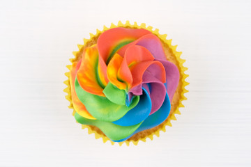 Cupcake with rainbow colorful cream in yellow cup on white wooden table. Top view.
