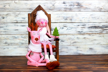 Doll-toy of a pink rabbit girl in a white hat with a green fir-tree and deer sitting on a chair. Love, romantic and friendship. Concept of joy and winter holidays