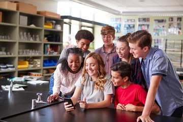 Group Of High School Students Looking At Message On Mobile Phone In Classroom