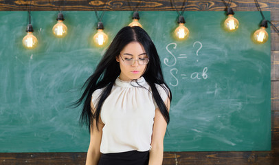 Lady strict teacher on calm face stands in front of chalkboard. Woman with long hair in white blouse stands in classroom. Teacher with glasses and waving hair looks attractive. Strict teacher concept.