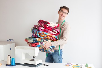 profession, business, hobby concept. there is a young tailor, who is standing in the light studio of desighn and sewing, he is holding lots of quilts made of bright patches and smiling happily