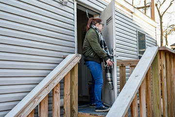 A woman walking outside the front door of her mobile home onto the front porch to turn and lock the door behind her wearing a dark green jacket and a neutral colored plaid scarf as she leaves.