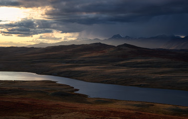 Fototapeta premium Colorful sunset above highland steppe shore of lake with dry yellow grass on the background of rocky mountains under storm sky