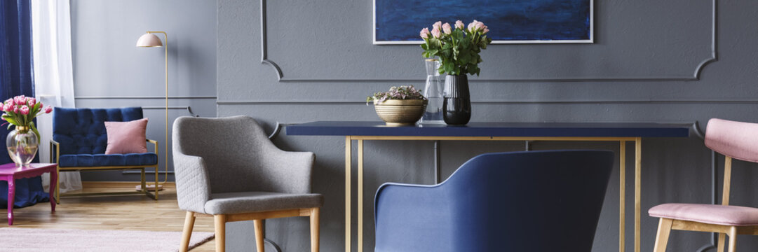 Chairs standing by the blue and gold dining table with pink roses in vase in dark grey open space living room interior