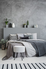 Striped carpet and stool in grey bedroom interior with sheets on bed with headboard. Real photo
