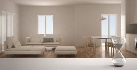 White table or shelf with crystal hourglass measuring the passing time over blurred empty space, living room with kitchen, architecture interior design, copy space background