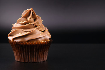 Cupcake with whipped chocolate cream, decorated, gold confectionery sprinkling on black background. Picture for a menu or a confectionery catalog.