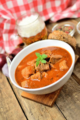 Close-up of pork goulash with pieces of meat in a bowl, garlic, pepper, onion, glass with cold beer and red checkered tablecloth in the background - vertical photo