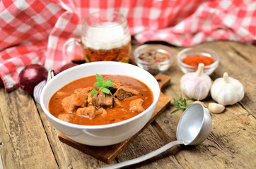 Pork goulash with pieces of meat in a bowl, garlic, pepper, onion, glass with cold beer, ladle and red checkered tablecloth in the background