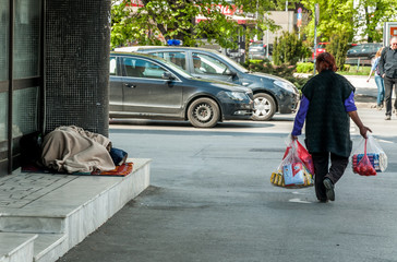 Homeless barefooted woman sleep on the urban street in the city on the sidewalk near the building while people are passing by, selective focus. 