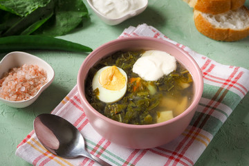 Green soup in a bowl. Sorrel soup with eggs and leaves, onion, bread on a green table. 