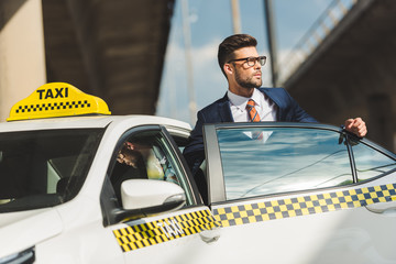 hansome young man in suit and eyeglasses looking away while sitting in taxi cab