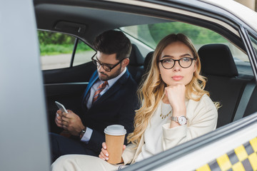 young woman holding paper cup and looking away while man using smartphone in taxi