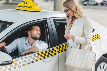 smiling blonde woman talking by smartphone and looking at male driver sitting in taxi