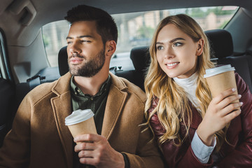 smiling stylish young couple with paper cups sitting together in taxi