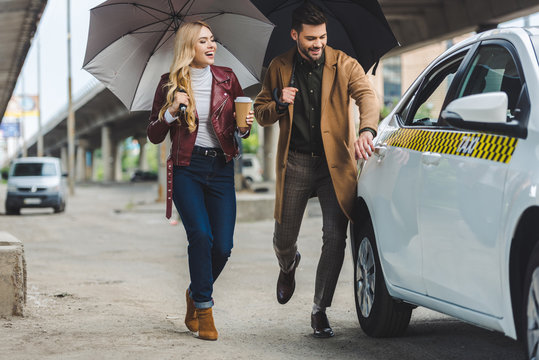 Smiling Young Couple With Umbrellas Running To Taxi Cab