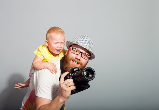 A photographer with a child in his arms takes an enthusiastic photograph against a gray background in the studio. The concept of "Children of Photography is not a hindrance"