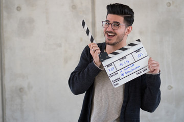 smiling young man holds a end slate slatterboard in front of a concrete wall with glasses // Junger...