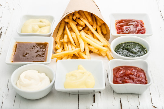 French fries wrapped in paper among different sauces in white saucers on the white wooden background with cracked paint. Top view.