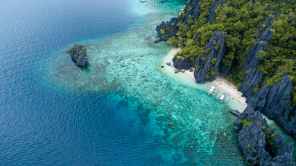 Fototapeta na wymiar Aerial drone view of boats above a tropical coral reef and small sandy beach surrounded by huge cliffs (Secret Lagoon, Miniloc)