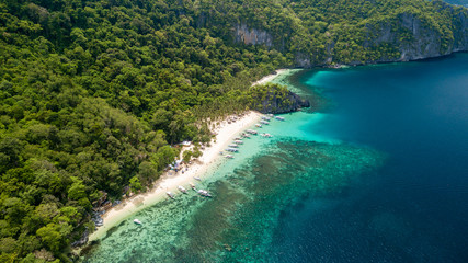 Aerial drone view of traditional Banca boats and coral reef surrounding a scenic tropical sandy beach (7 Commando Beach, El Nido)