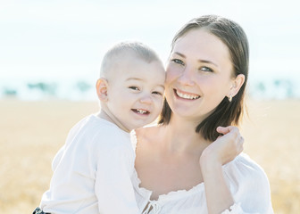 Fototapeta premium Family young happy pretty mother holding little baby son and smiling in golden wheat or rye field at sunny summer day. Blurred skyline background. Concept simple rustic life childhood and motherhood