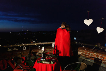 Boy and girl hug each other tender standing on the rooftop in the rays of evening lights and having a romantic dinner
