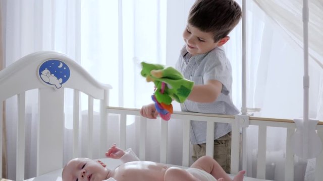 brotherly love, kid with toy in arms is entertaining with infant in cot near brightly lit window