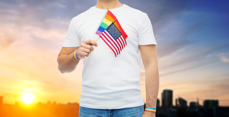 lgbt, same-sex relationships and homosexual concept - close up of man wearing gay pride rainbow awareness wristbands and holding american flag over city sunset background