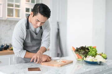 Papier Peint photo autocollant Cuisinier Homme asiatique confus lisant sa tablette avec un regard pensif et pensif tout en se tenant dans sa cuisine tout en cuisinant et en préparant un repas
