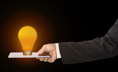 Businessman hand holding mobile phone and stack of money coins with light bulb ideas on top; concept of financial planning and technology