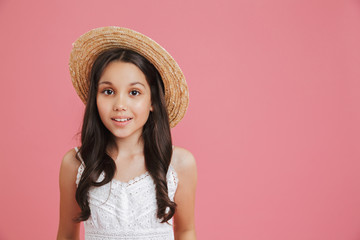 Portrait of cute summer girl 8-10 wearing white dress and straw hat looking at camera with smile near copyspace, isolated over pink background