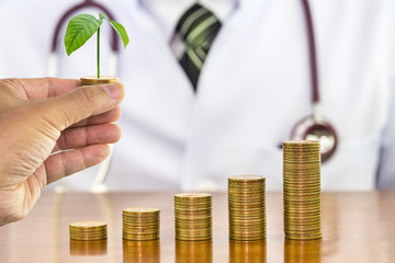 Man hand holding money coin with tree growing and blurry the doctor stethoscope around neck a scene in the back, concept of financial health with medical expenses and saving money
