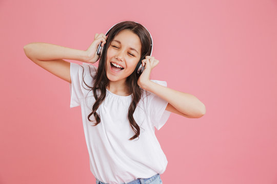 Happy teen girl 8-10 in casual clothing singing with closed eyes while listening to music via wireless headphones, isolated over pink background