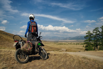 Motorcycle traveler man in helmet with suitcases standing on extreme rocky road in a mountain valley in cloudy weather on the background of endless steppe
