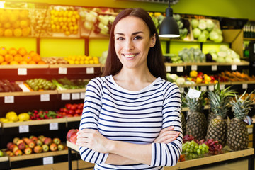 Attractive healthy smiling woman in casual clothes looks at the camera against the background of shelves in a supermarket. Fruits and vegetables are great.