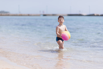 Fototapeta na wymiar Adorable boy standing in the water on the beach playing with a ball. Family vacation or holiday concept.