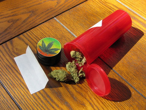Marijuana buds in a prescription container with a grinder and rolling paper for a joint on a wooden table, the strain of marijuana is Green Crack 