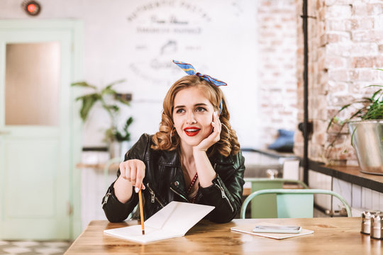 Young lady in leather jacket sitting at the table with notebook and pencil in hand and joyfully looking aside while spending time in cafe