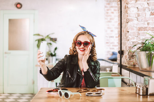 Beautiful smiling lady in leather jacket sitting with many different sunglasses on table and joyfully taking photos on cellphone while spending time in cafe