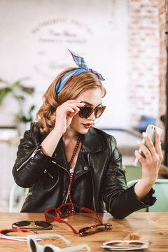 Young lady in leather jacket sitting with many different sunglasses on table and taking photos on cellphone while spending time in cafe