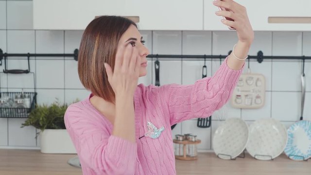A nice woman is holding a smartphone, making selfie in the kitchen. A modern woman blogger is taking pictures of herself on the phone. Lifestyle photo content.