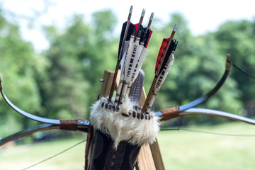 Medieval set of old wooden arrows in leather case and bows hanging on stand. Green forest blurred...