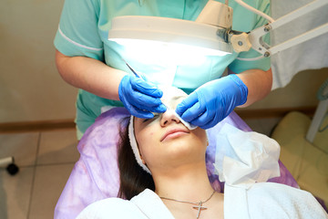 Cosmetologist masseuse cleaning woman face at spa in cosmetic cabinet under a lamp. Clean fresh skin, wearing white bathrobe and hair bandage, doing cosmetic procedure, spoon face cleaning.